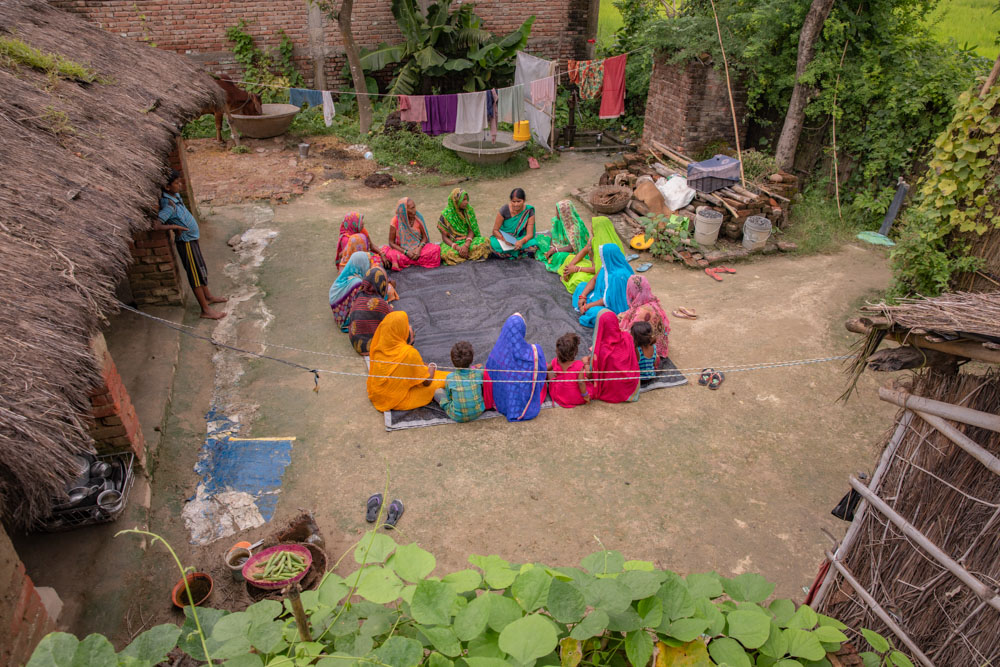 A group of women sitting in a circle in the lawn in front of a house in a village, and talking with each other.