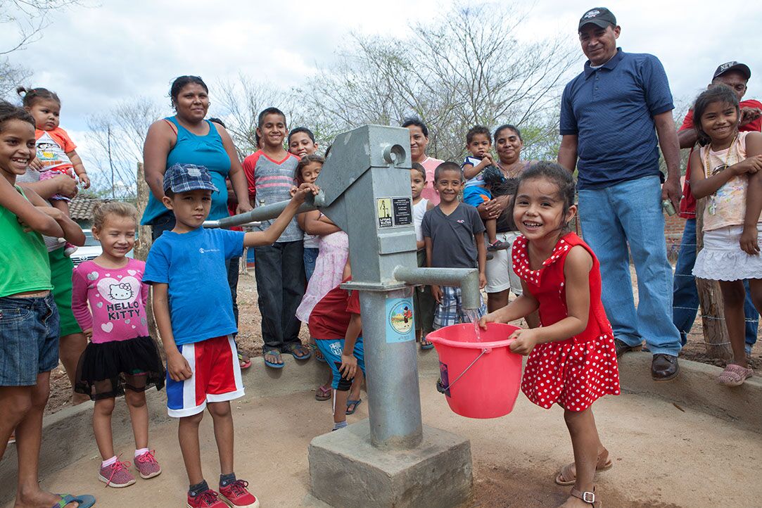 Little girl holding a bucket under a water spicket and surrounded by the people in her town.