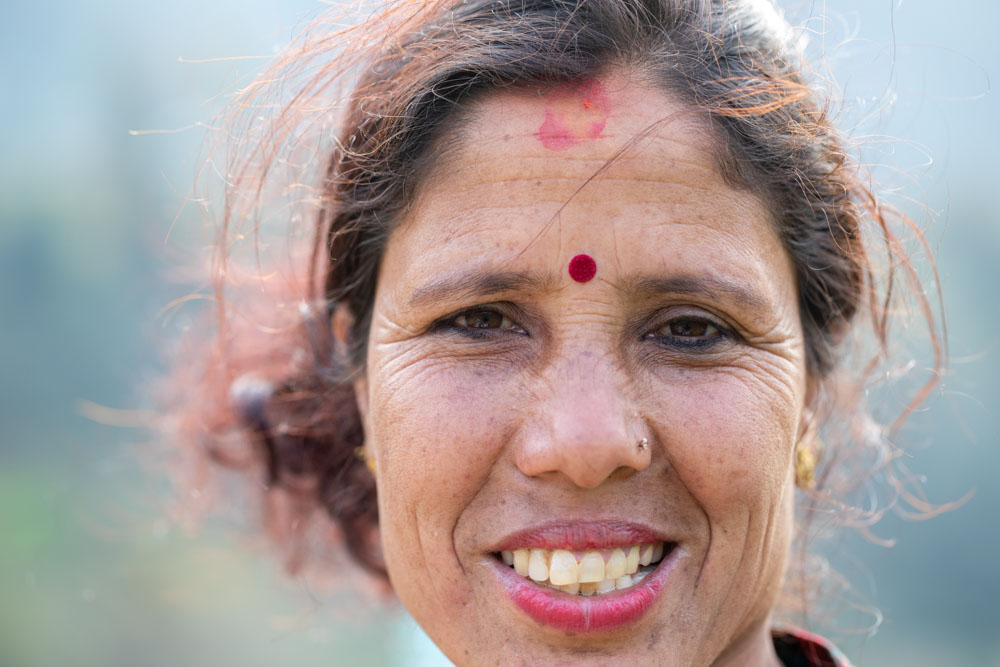 A close-up portrait of a smiling woman with a red bindi on her forehead.