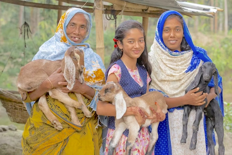 Two women in ornamental gowns, standing on either side of a young girl. All are holding baby goats and smiling.