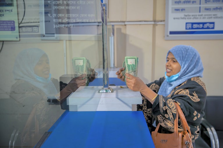 A woman wearing a blue headscarf and embroidered black dress conducts a financial transaction at a service counter, holding currency while reflected in the glass partition that separates her from the teller.
