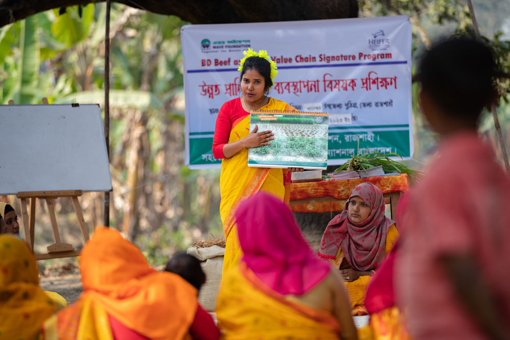 A woman in a vibrant yellow saree conducts a training session outdoors, holding up a chart, with women in colorful attire listening attentively.
