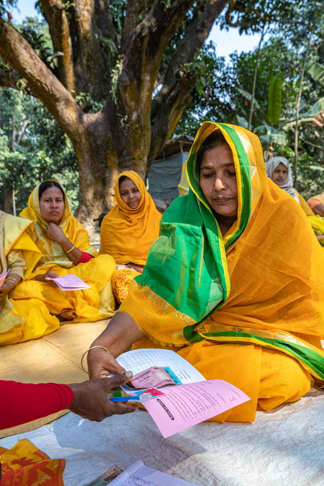 A woman in a yellow and green saree sits on the ground, reading a pamphlet, with other women in yellow sarees in the background.