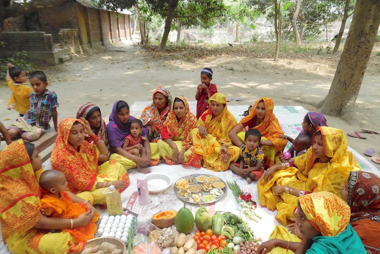 A community and their children gathered together around their harvest of many fruits and vegetables.