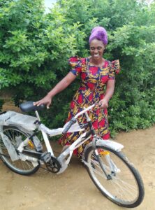 A smiling woman wearing a purple headscarf and floral dress admires her new bicycle.