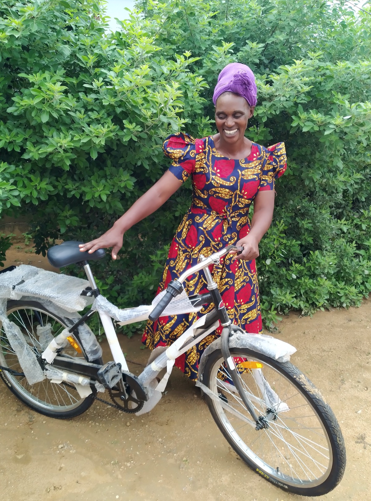 A smiling woman wearing a purple headscarf and floral dress admires her new bicycle.