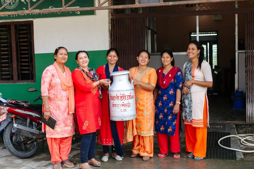 Six Nepali women stand next to each other, smiling, and holding a large can of milk.
