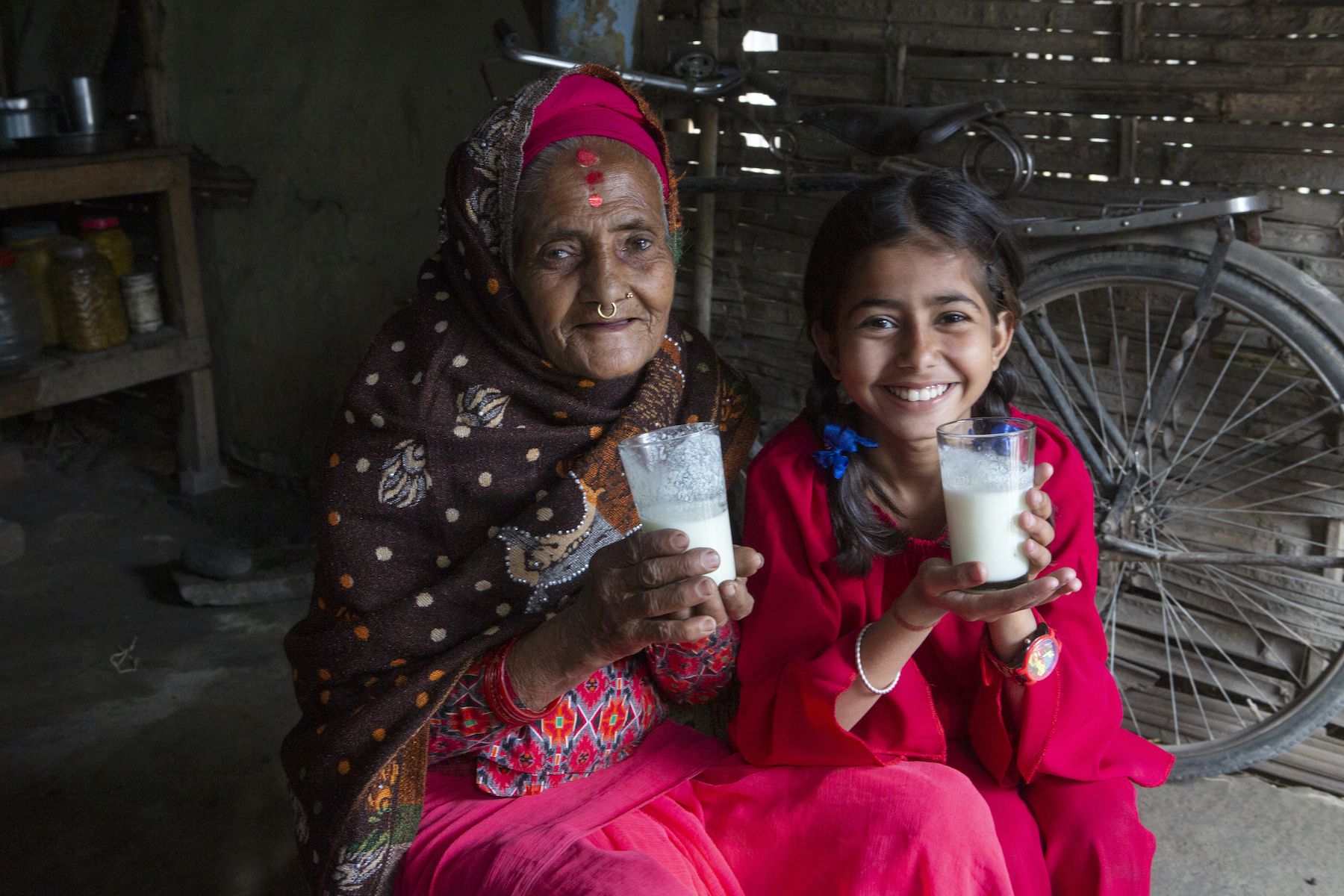 A smiling Nepali woman in her 70s and her 12-year-old granddaughter each hold a glass of milk in their hands.