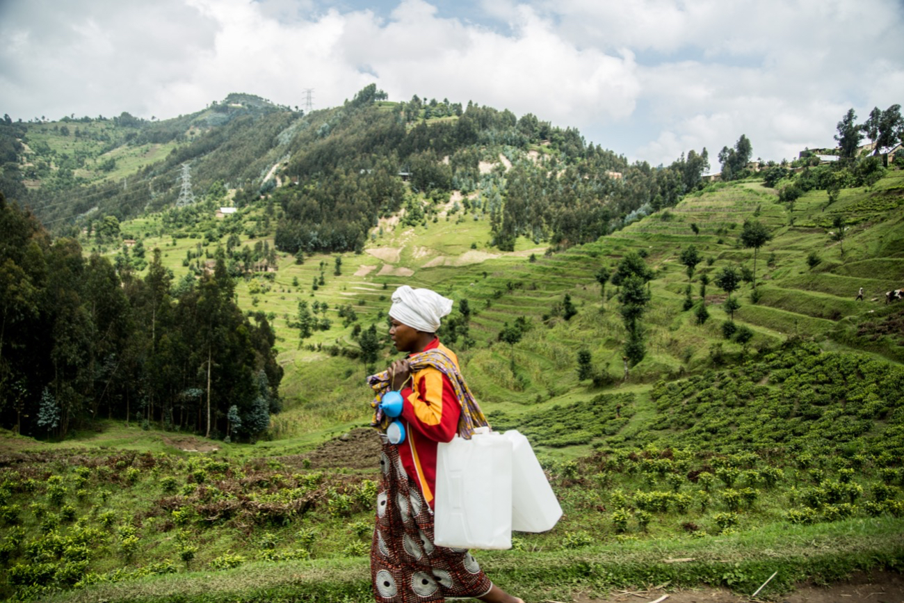 A woman walks through a green hillside with containers over her shoulder.