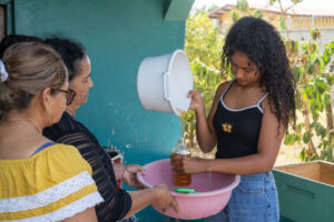 A young Honduran woman pours honey from a white bucket into a plastic bottle while two other women watch.
