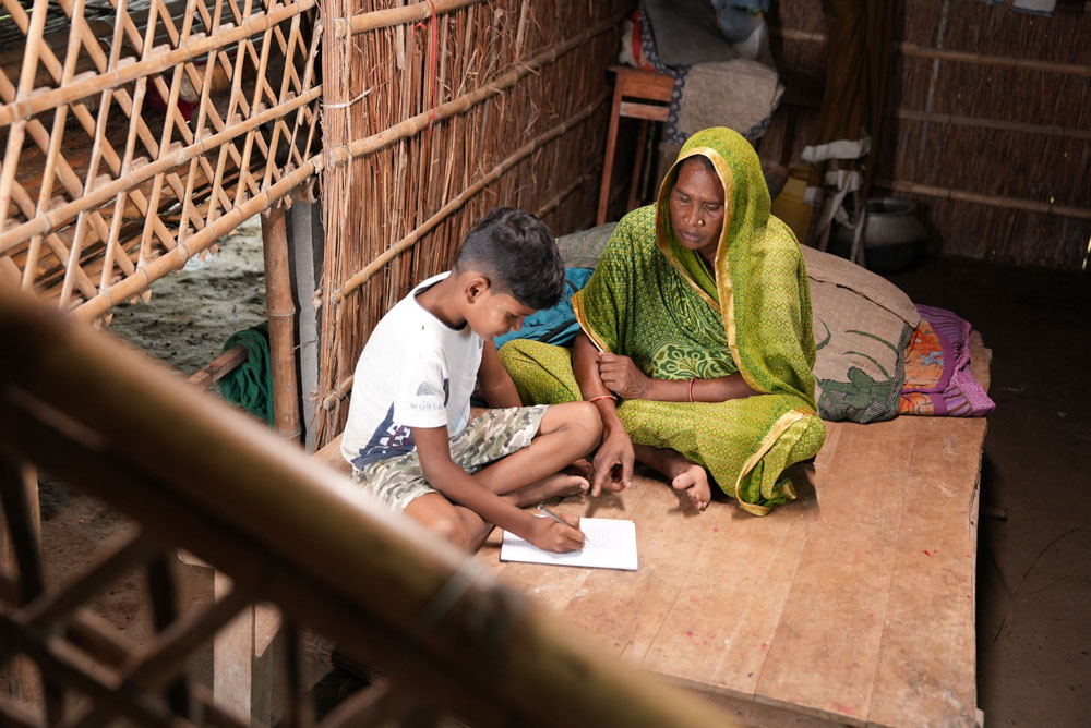 An Indian woman wearing a green sari sits next to a boy with schoolbooks on the floor in front of him.