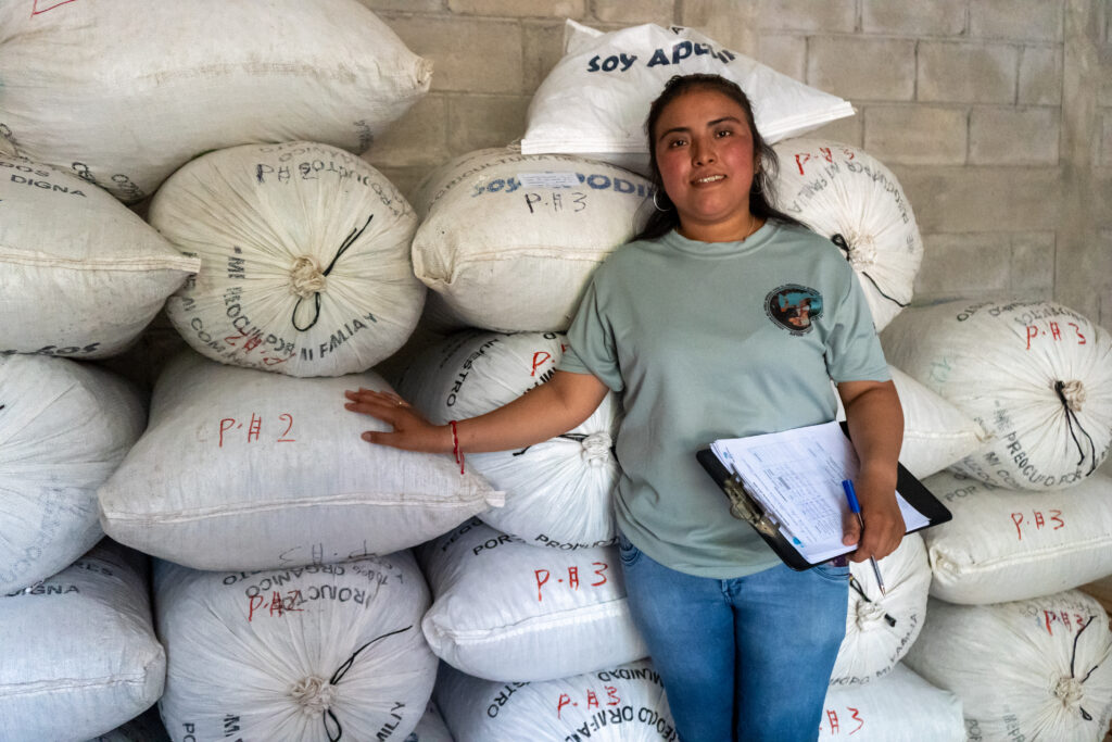 A woman stands beside stacked sacks of cacao.