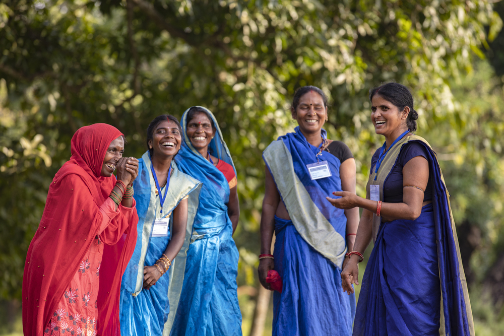 Five Indian woman wearing colorful clothing stand together, laughing.