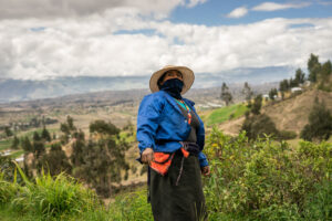 A person in a straw hat, blue jacket, and face covering stands on a hillside overlooking a vast rural valley beneath a dramatic cloudy sky.