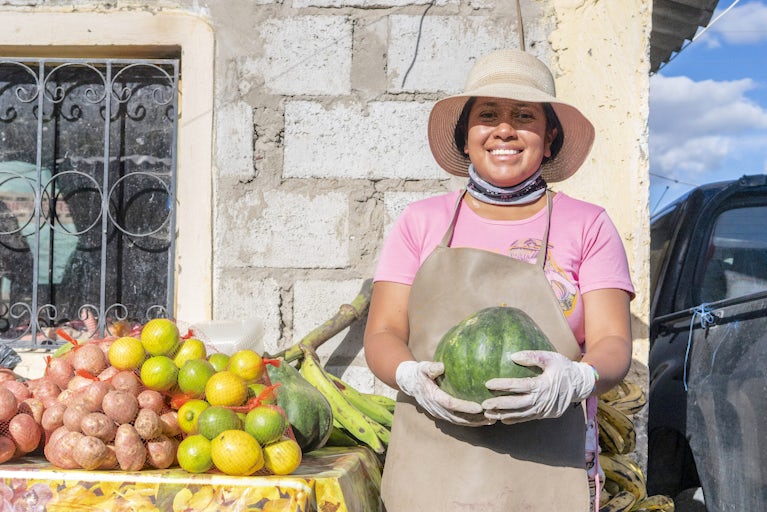 Woman wearing a hat and holding a watermelon while she stands next to table of fruit.