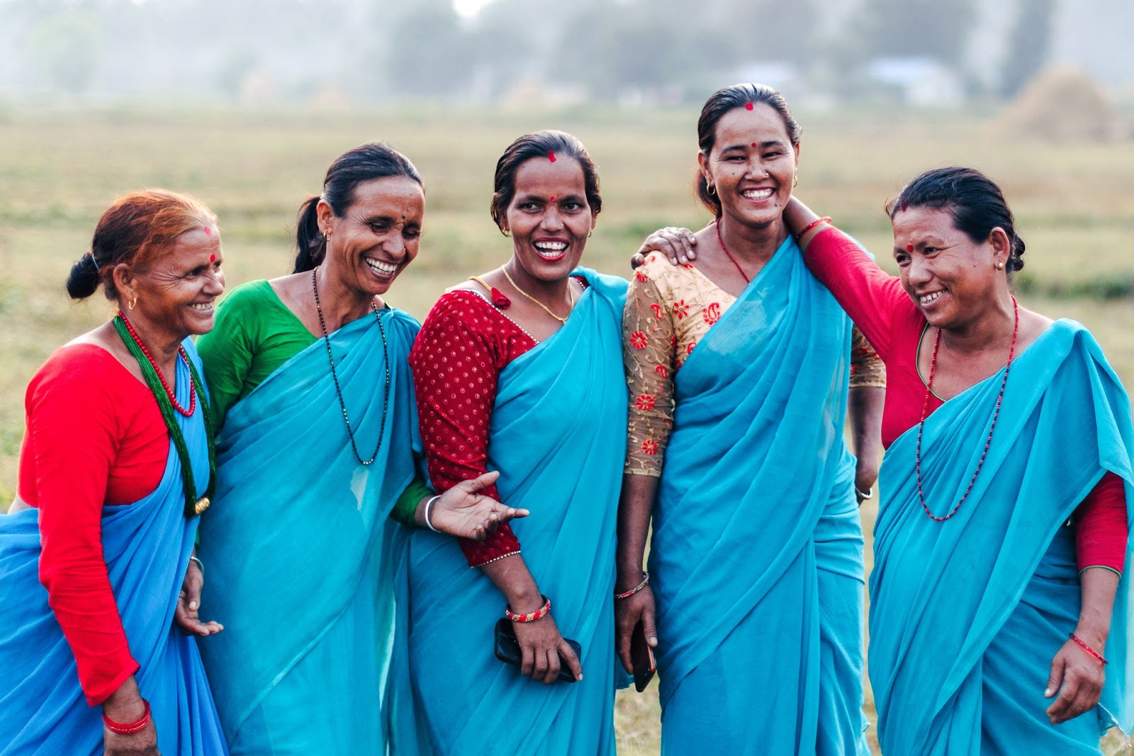 Five women wearing blue sarees and red blouses stand together outdoors, smiling and laughing. They appear to be enjoying each other’s company in a rural setting with green fields in the background.