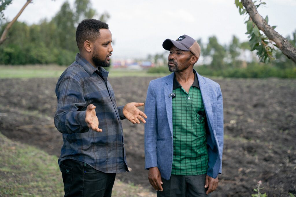 Two men stand in a farm field talking.