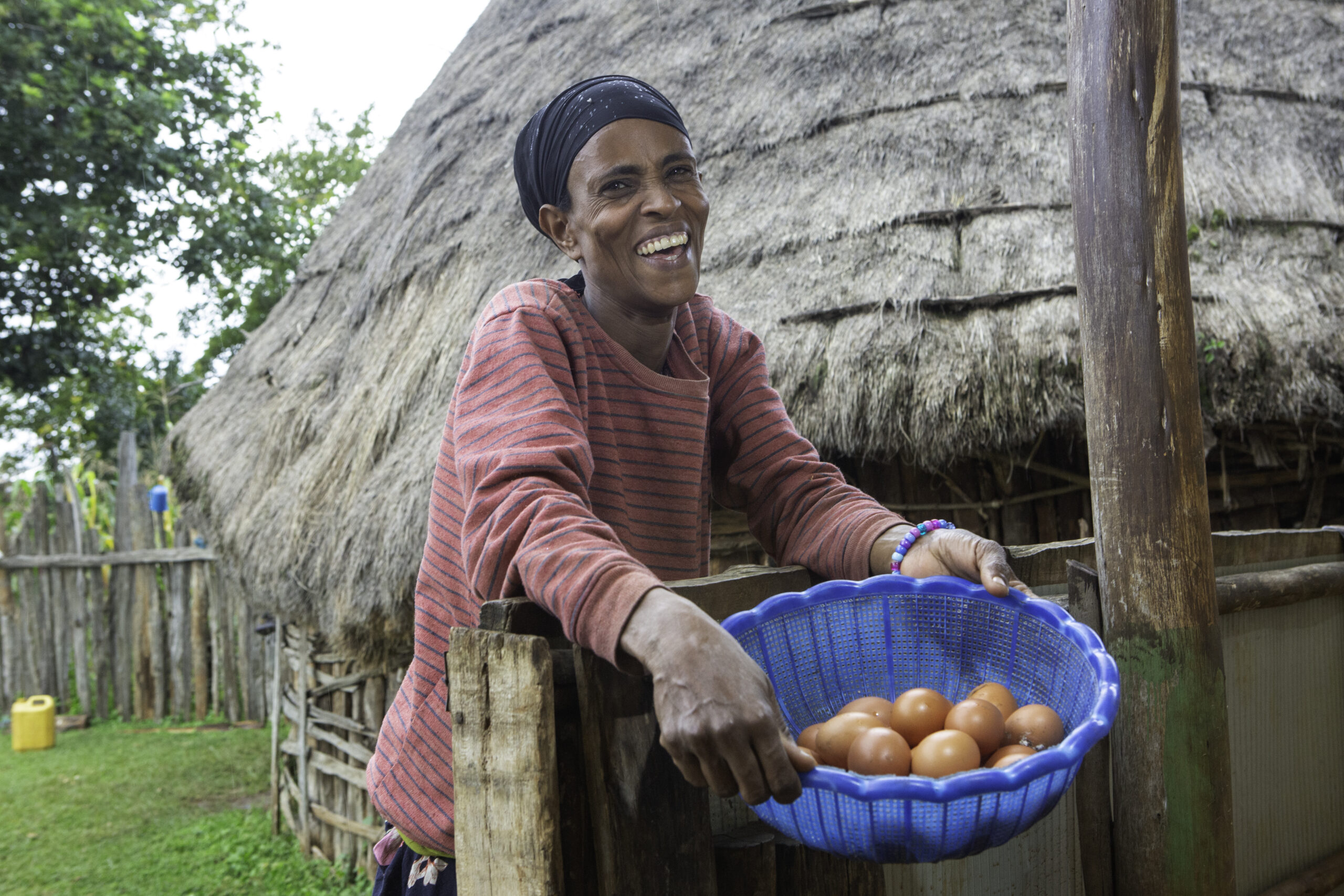 A woman smiles while holding a blue basket filled with brown eggs outside her chicken coop.