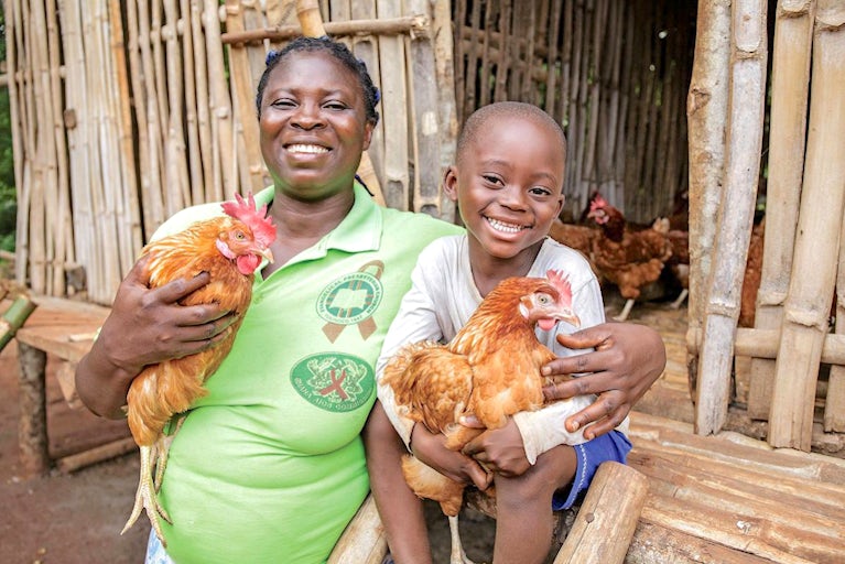 Woman and child smile while holding chickens in front of wooden structure.