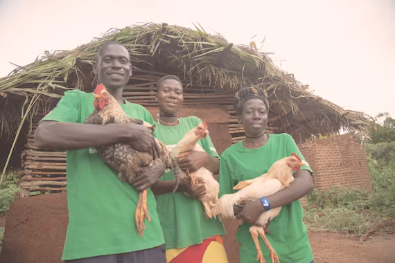 Three people in green shirts holding chickens in front of a thatched hut.