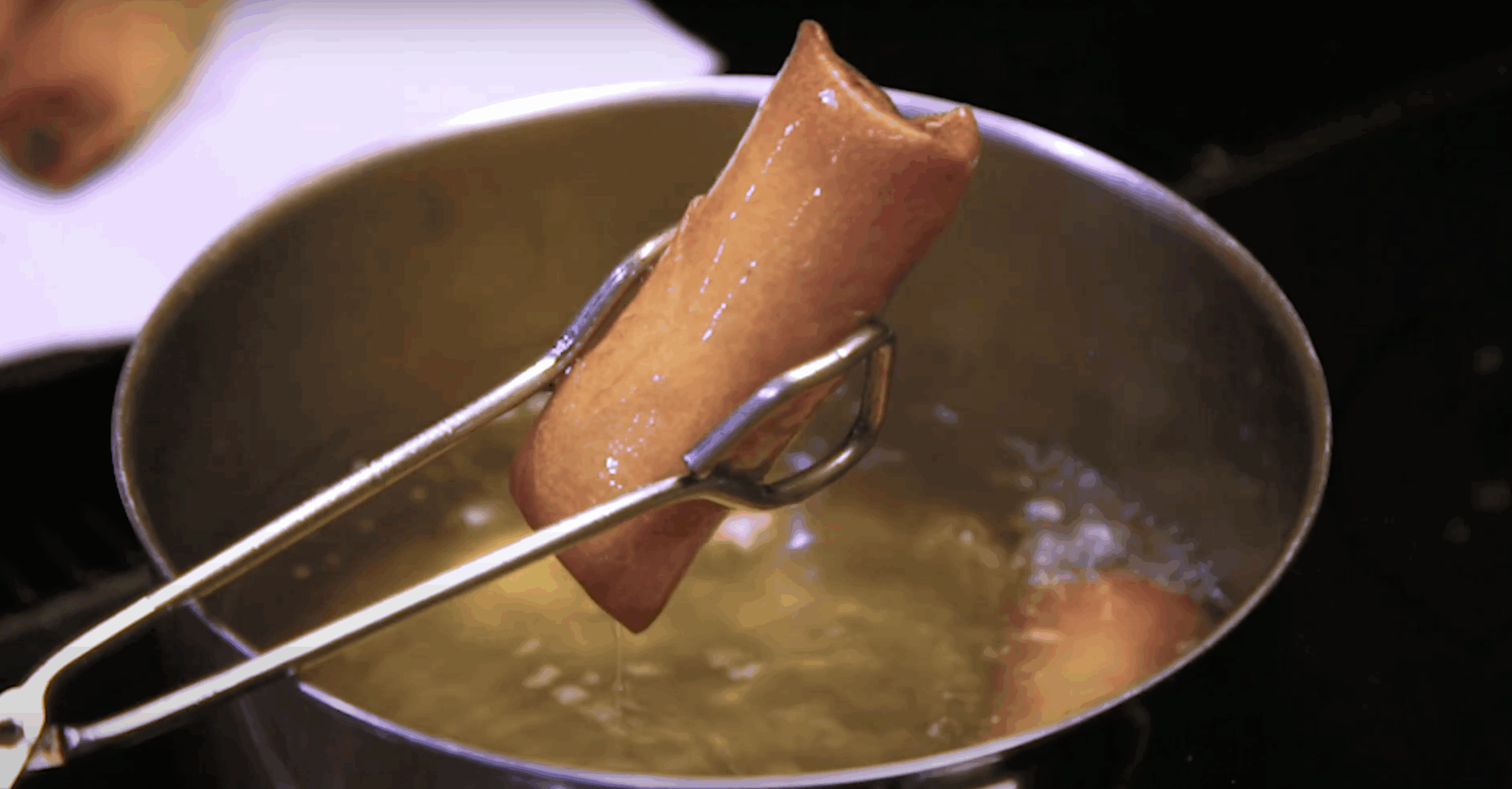 A pair of tongs remove a golden brown spring roll from hot oil.