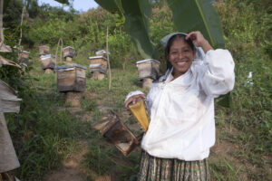 A smiling Guatemalan woman in a bee suit poses with her beehivevs.