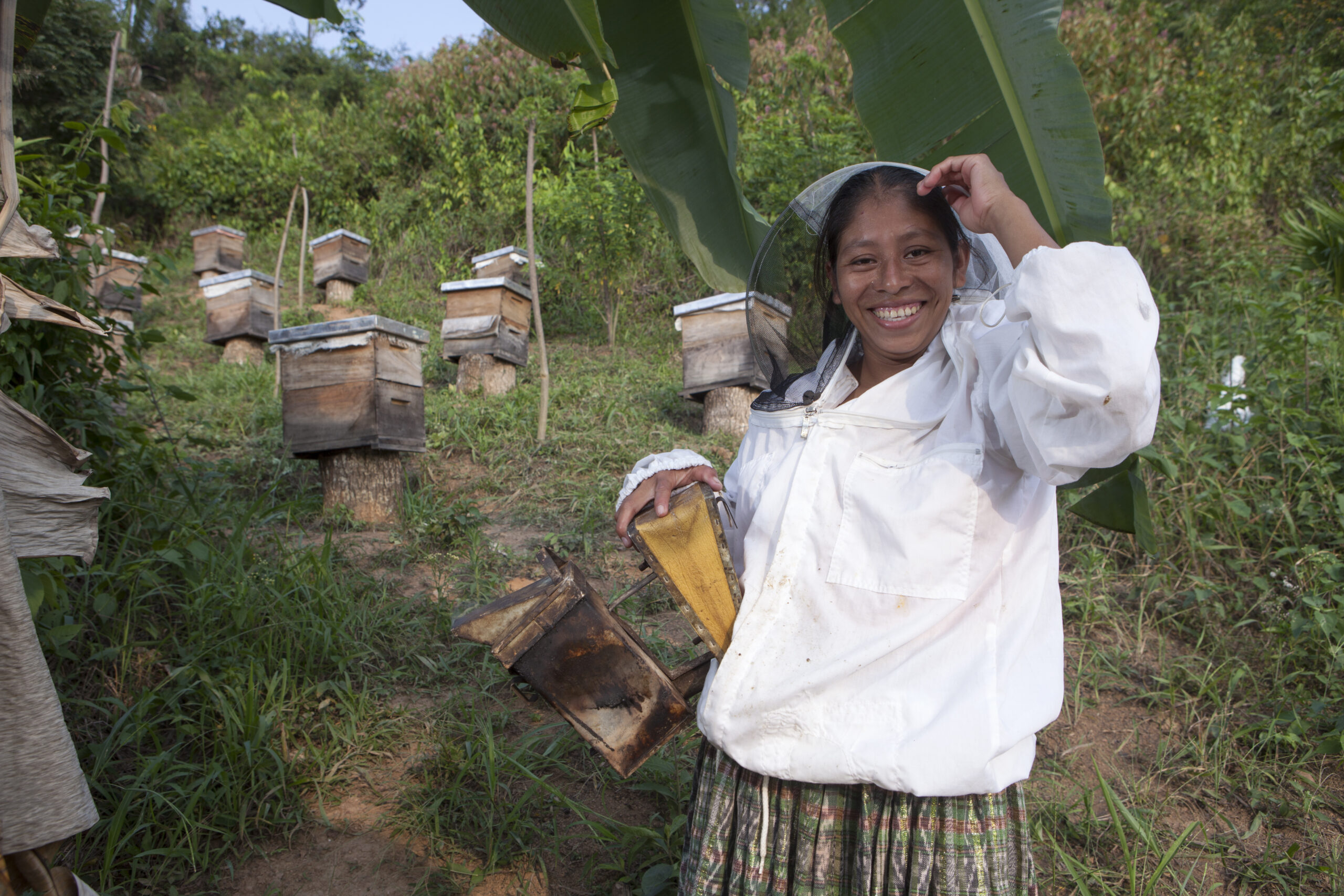 A smiling Guatemalan woman in a bee suit poses with her beehivevs.