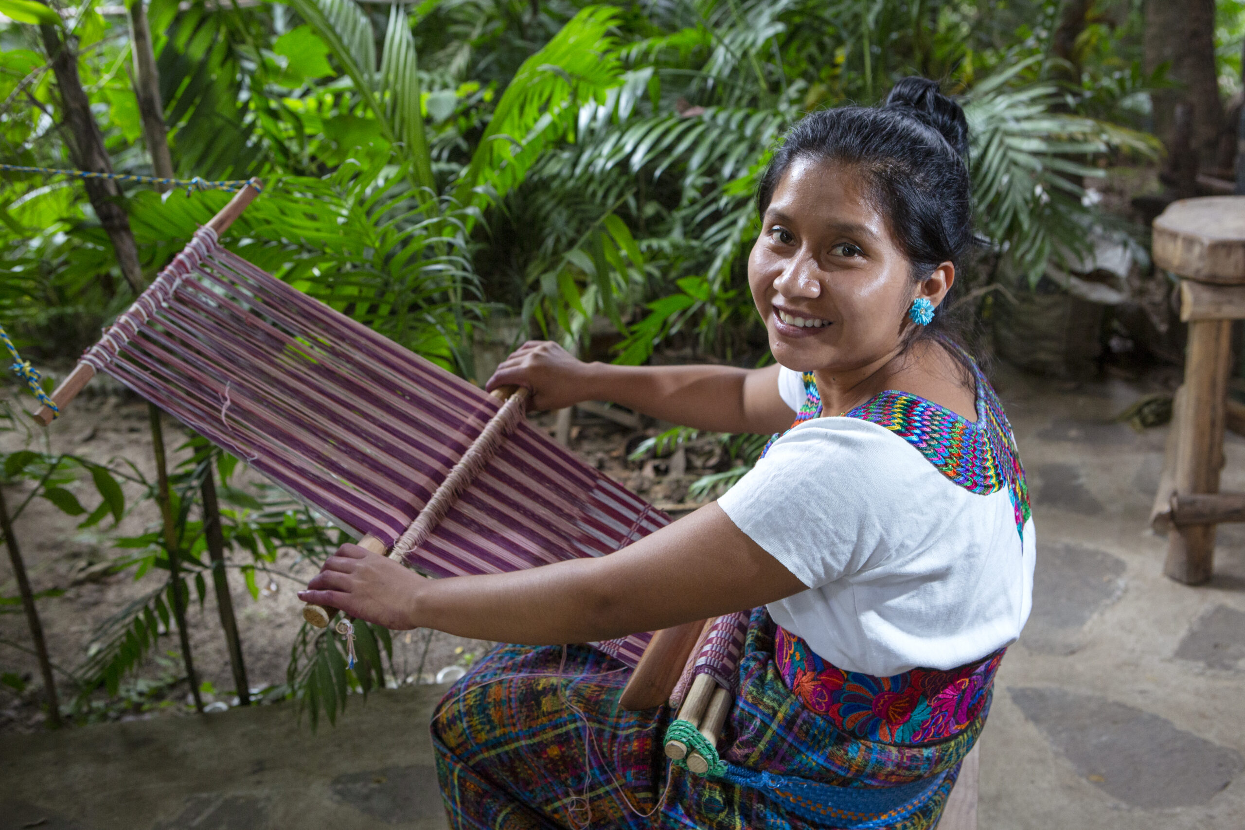 A woman sits at a backstrap loom weaving a shawl with various shades of crimson. She is smiling at the camera. 