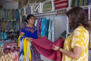 Two women (one dressed in a blue blouse and the other in a yellow blouse) look over a red shawl