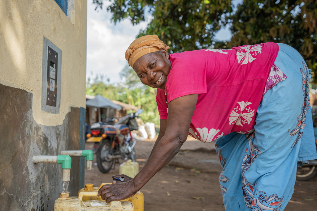 A woman smiling as she fills yellow jerry cans at a community water tap beside her home.