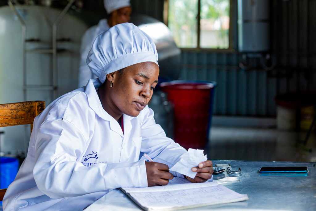 A woman in a lab coat writes notes while reviewing a sample in a processing facility.