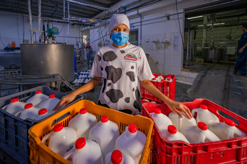 A woman wearing a blue medical mask standing in a factory behind two crates filled with gallon jugs of milk.