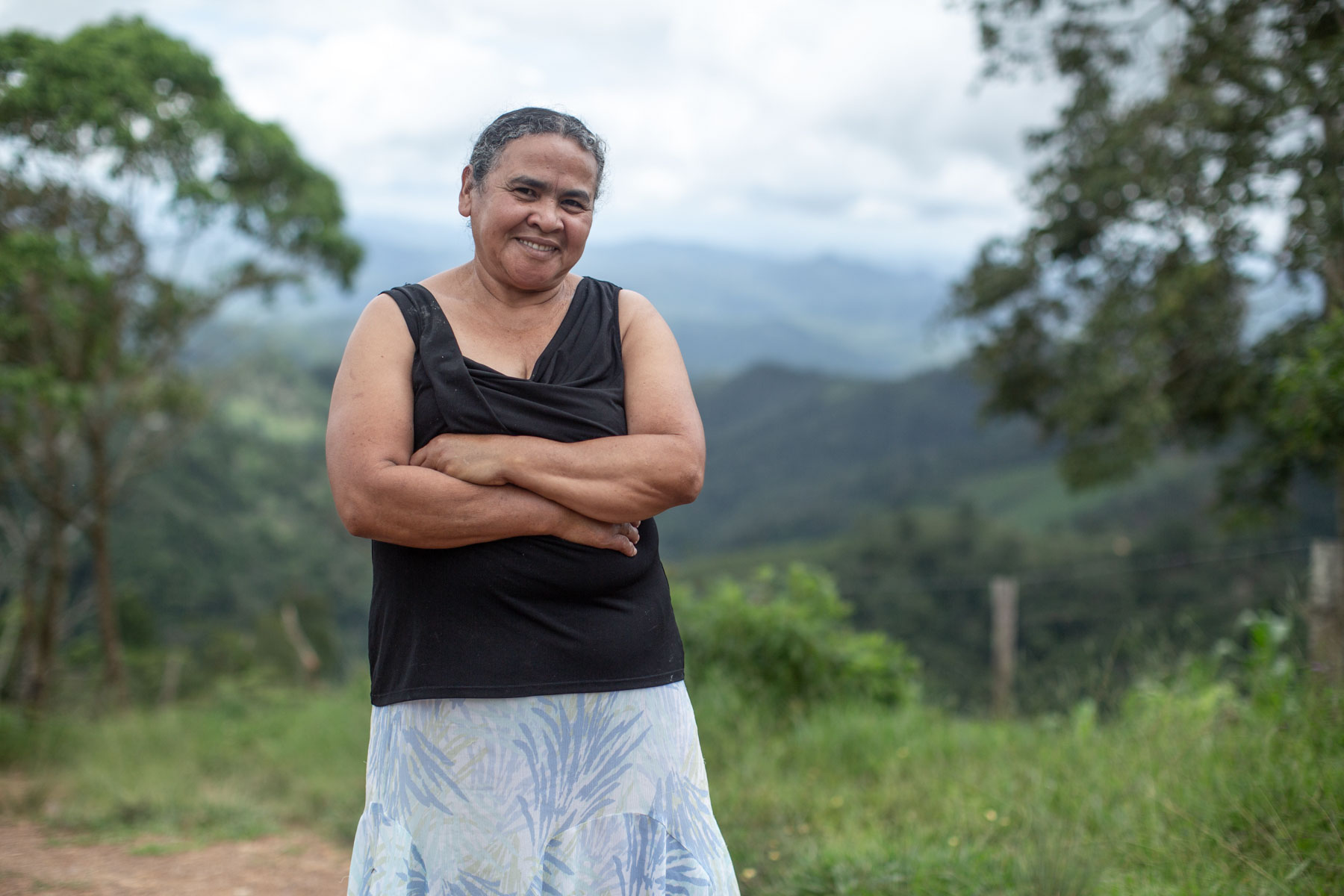 A smiling woman stands with her arms crossed in a green landscape.