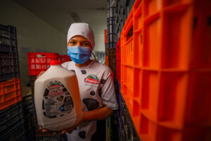 A masked worker in a dairy uniform holds a gallon of milk in a storage room surrounded by colorful crates.