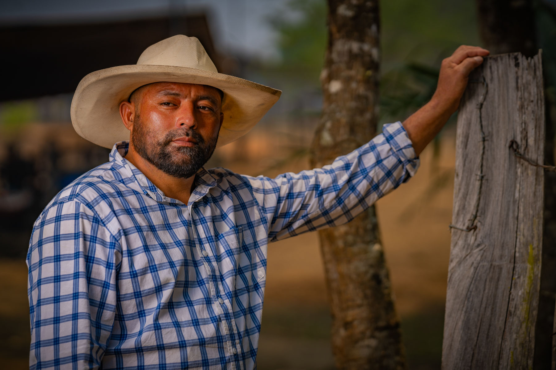 A man in a cowboy hat and plaid shirt stands by a wooden post on a farm.