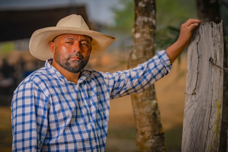 A man in a cowboy hat and plaid shirt stands by a wooden post on a farm.
