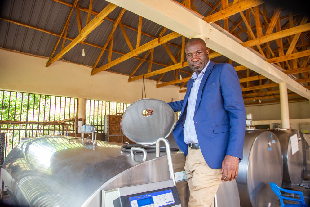 A man in a navy blue suit leans on a large, metallic milk cooling tank inside a dairy facility.