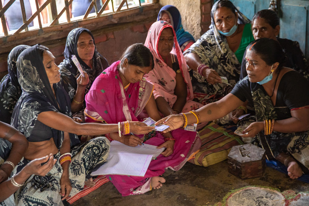 A group of women, some in saris, sit in a circle on the floor indoors, engaging in a financial transaction with cash in hand.