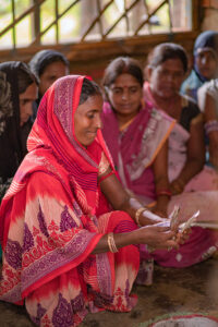 A woman in a red and pink saree counts money, focusing intently, with other women seated around her in a semi-circle.