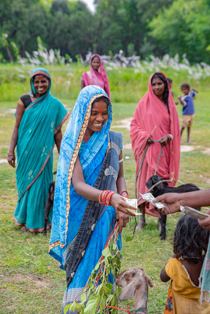 A woman in a blue saree exchanges money in a rural setting with goats and other villagers around her.
