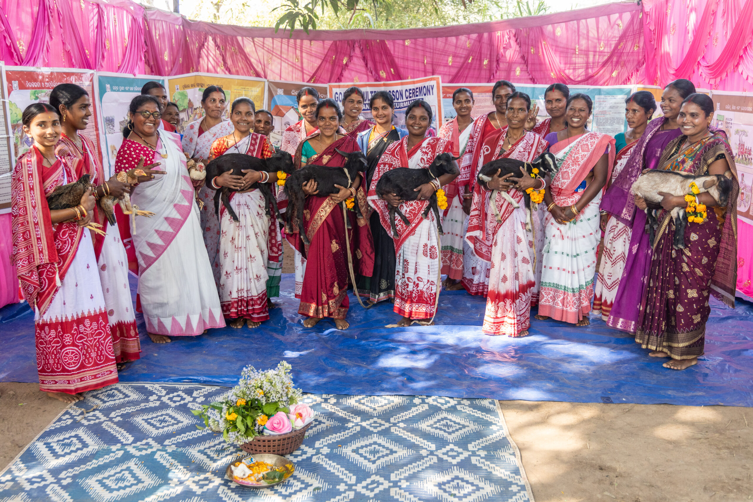 A group of women farmers in colorful saris stand together holding goats and chickens.