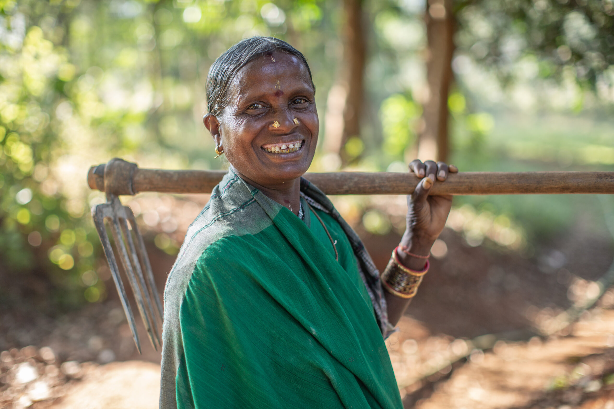 A woman farmer in a green sari stands outdoors and carries a hand tool over her shoulder