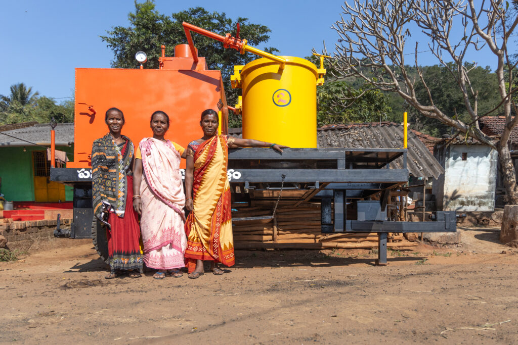 Three women stand beside turmeric processing equipment.