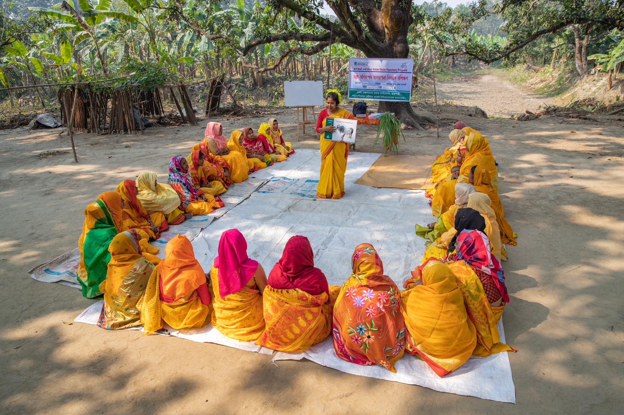 A group of women sitting in a semi-circle and a community trainer standing at one end conducting training with the help of a booklet.