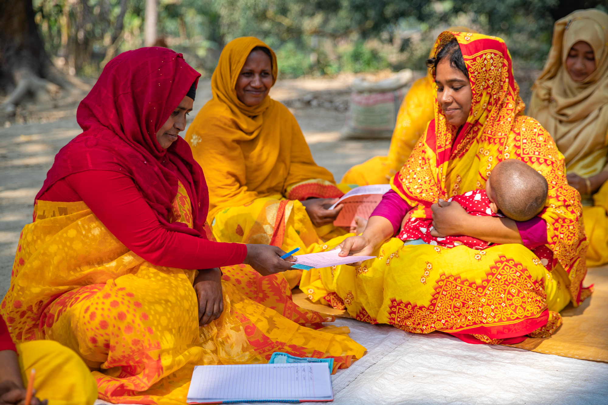 A woman holding an infant receiving her savings ledger back after it has been filled by her group member.