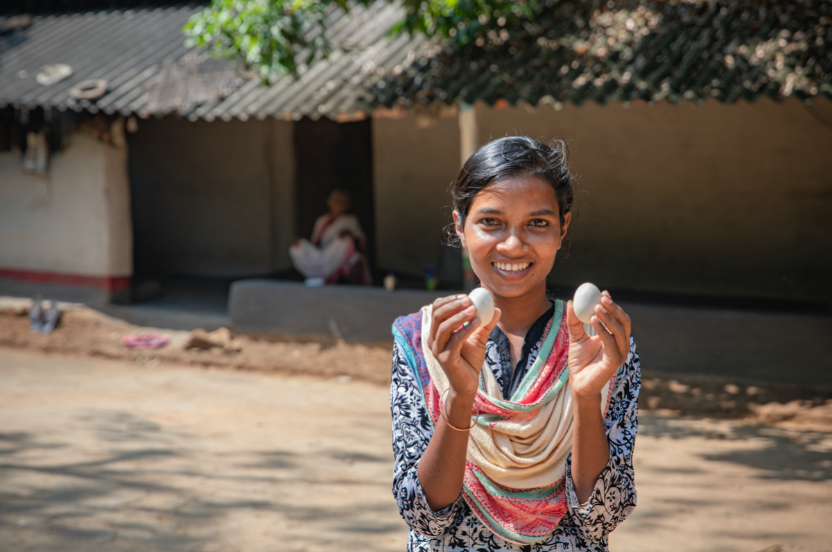 A young Indian woman holds a chicken egg in each hand and smiles at the camera.