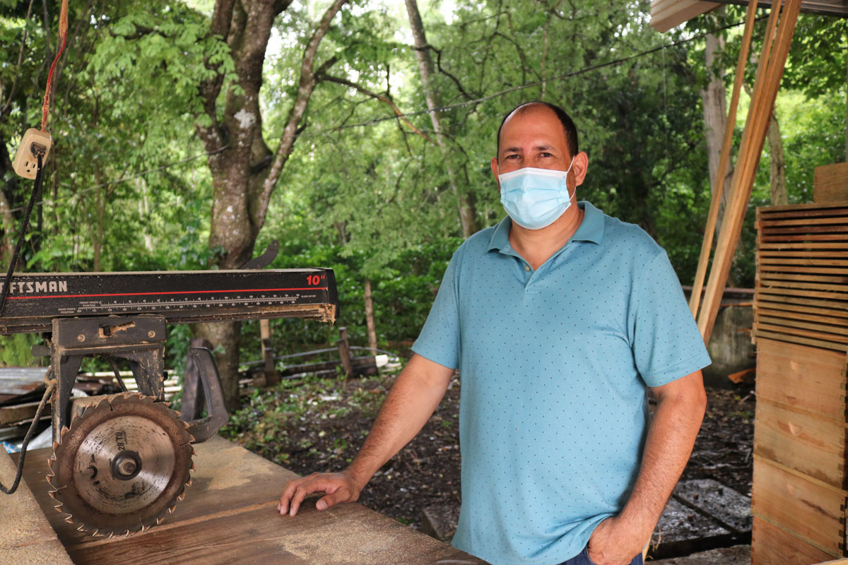 A Honduran man wearing a mask and blue shirt stands by his carpentry workshop.