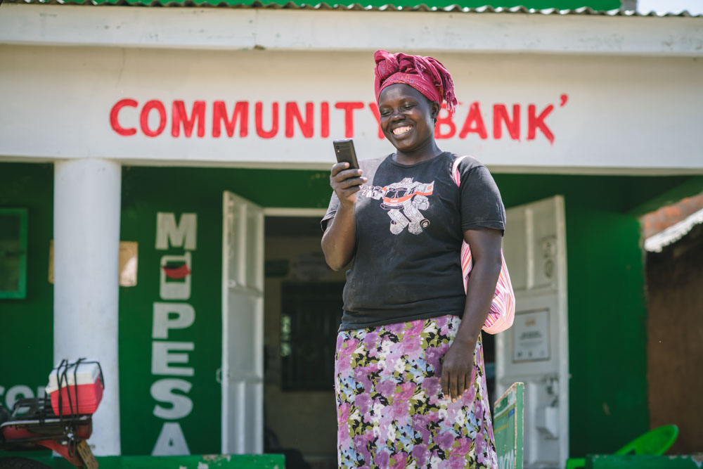 A woman in a printed skirt and red headwrap stands outside a community bank holding a mobile phone.