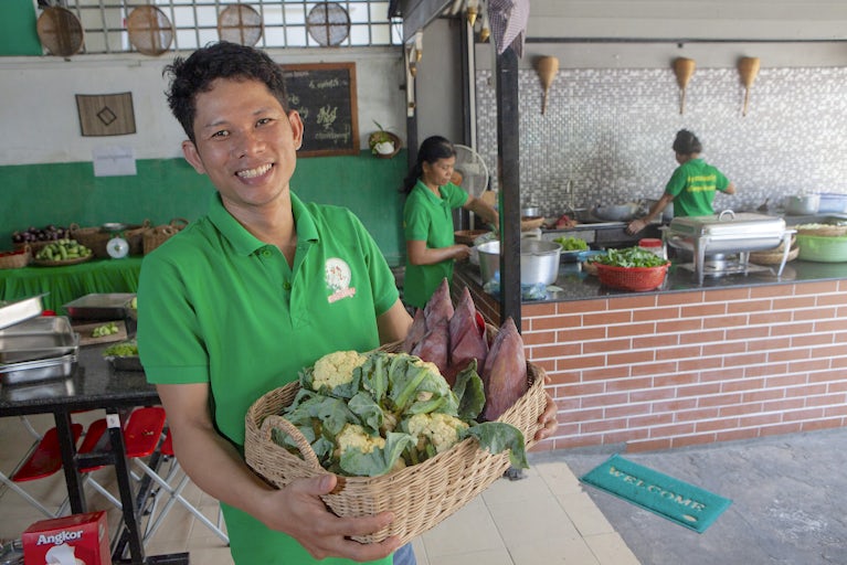 Smiling man in green shirt holds basket of fresh vegetables in commercial kitchen.