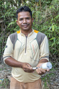 A man holds a syringe and vaccine vial while standing in a rural setting.
